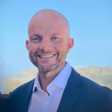 Kenneth Bonnici, smiling, in a dark jacket and open-necked white shirt, photographed outdoors against a clear sky and a low ridge of Maltese countryside.